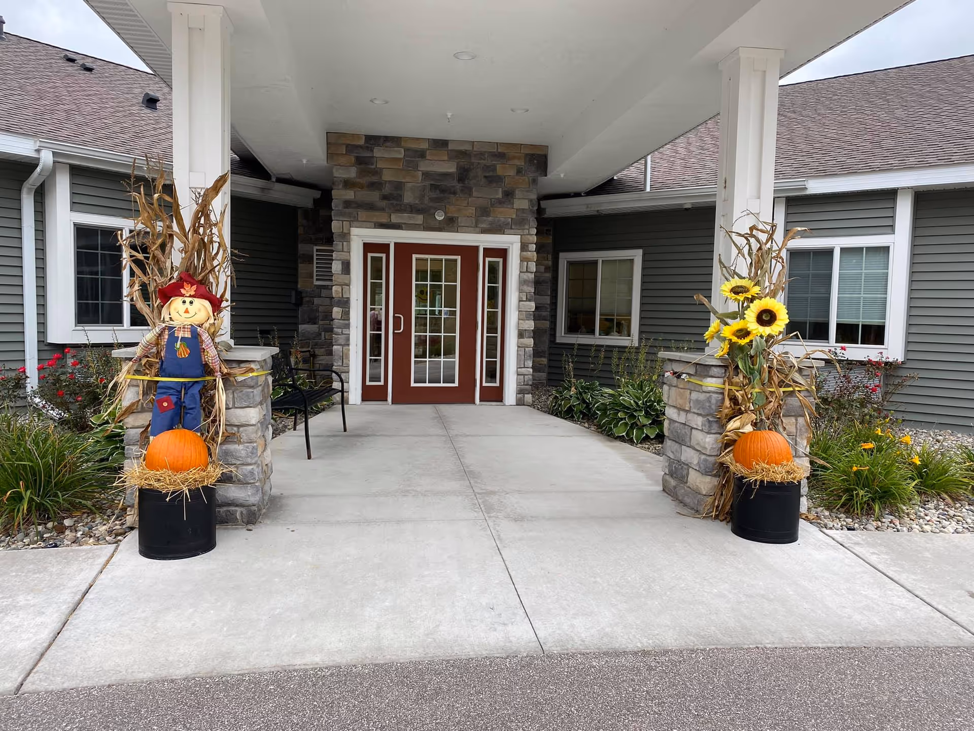 Entrance to a building with a covered walkway supported by white pillars. The entrance has double glass doors framed with stone. On either side of the walkway, there are autumn decorations including pumpkins, cornstalks, a scarecrow on the left, and sunflowers on the right. The building exterior is gray with white trim and windows.