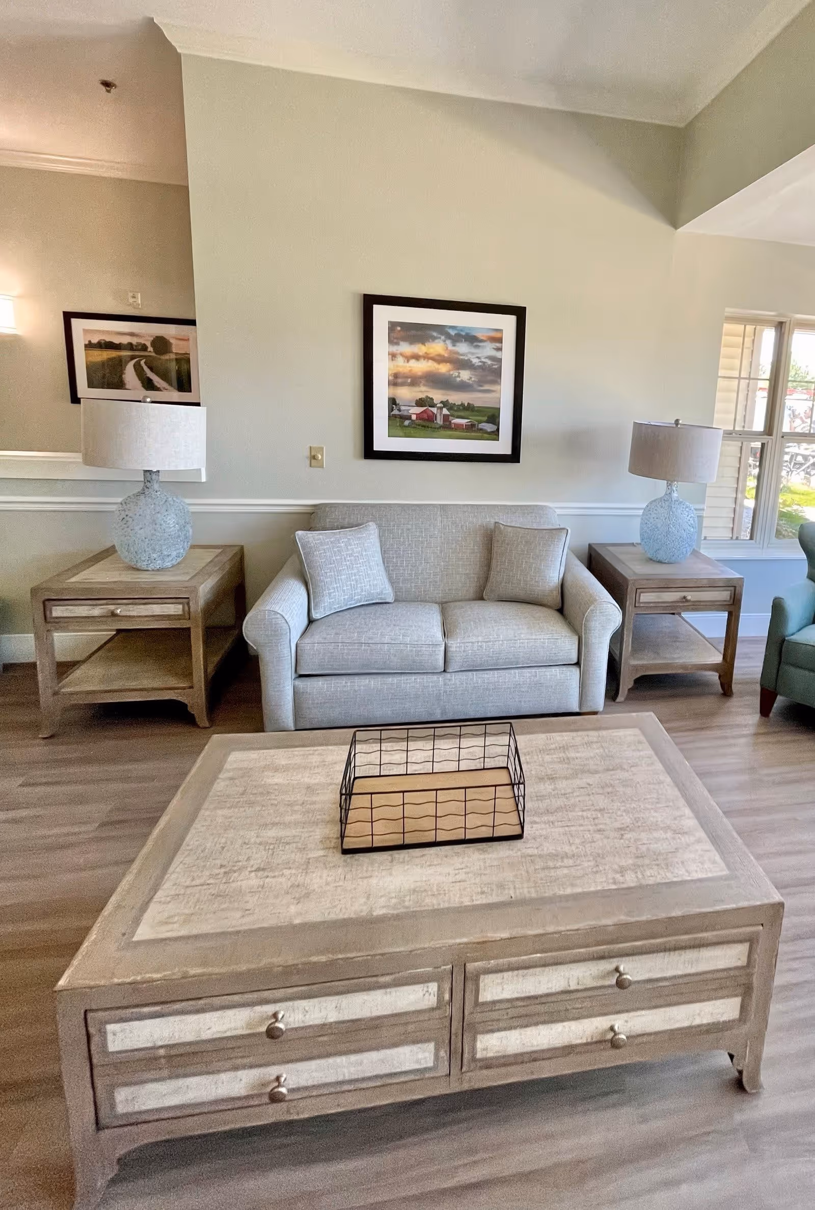 A cozy living room area featuring a light gray loveseat with two matching pillows, flanked by two wooden side tables each holding a textured blue lamp. A large wooden coffee table with drawers and a decorative wire basket sits in front of the loveseat. The walls are light-colored with a framed landscape painting above the loveseat and a window to the right letting in natural light.