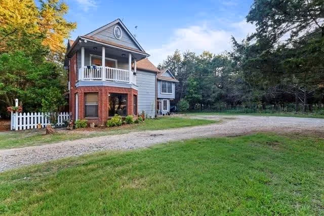 Two-story brick and siding home with a front balcony, gravel driveway and grassy yard surrounded by trees.