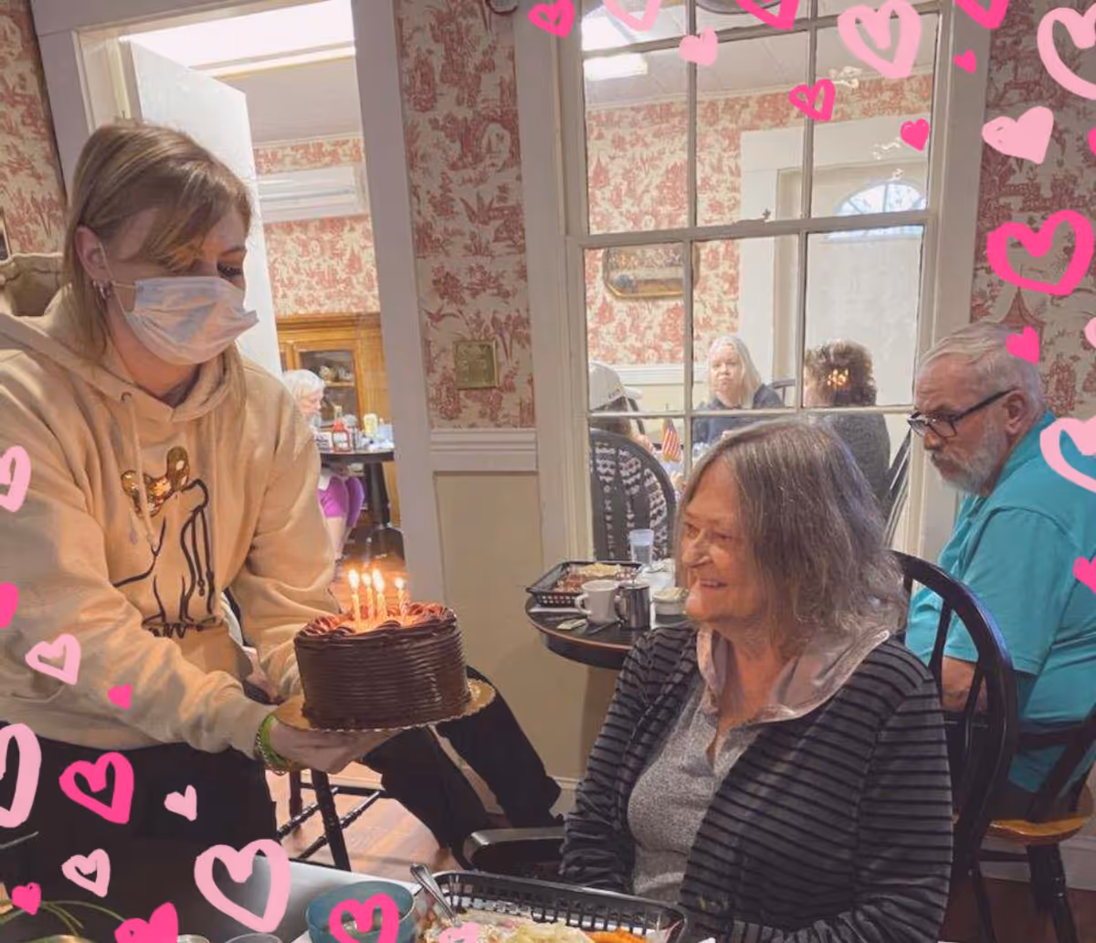 A woman wearing a face mask and a beige hoodie is presenting a chocolate birthday cake with lit candles to an elderly woman seated at a table in a dining area. The elderly woman is smiling and wearing a striped cardigan. In the background, other elderly people are seated at tables, and the room has floral wallpaper and a window with multiple panes. Pink heart graphics are overlaid on the image.
