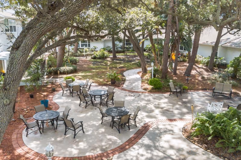 Outdoor patio area with several round tables and chairs arranged on a concrete surface bordered by red bricks. The patio is surrounded by trees and greenery, with a winding pathway leading through a garden area. White buildings are visible in the background.