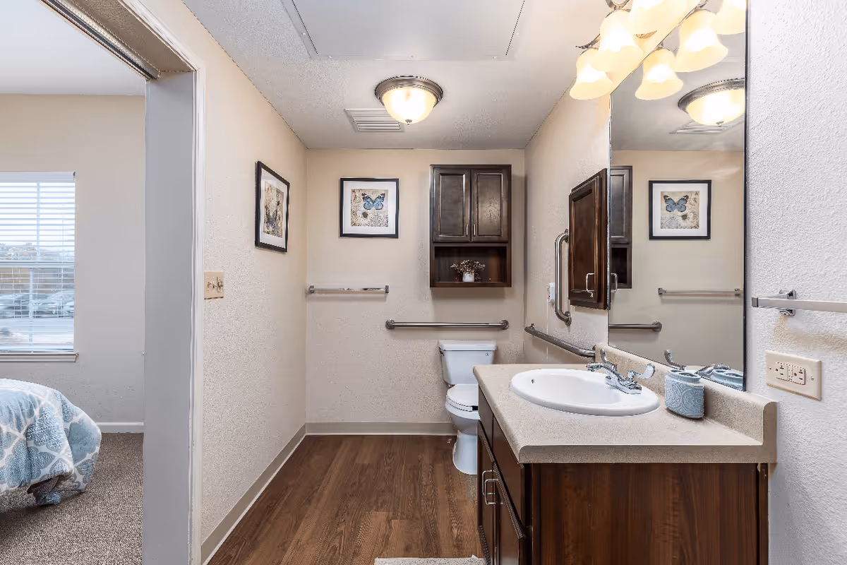 A bathroom with a wooden floor and beige walls featuring a white toilet with grab bars on the wall behind and beside it. Above the toilet is a dark wooden cabinet with a small plant. To the right is a countertop with a white sink, a soap dispenser, and a large mirror above it. The bathroom is well-lit with ceiling and wall lights. On the left side, there is an open doorway leading to a bedroom with a bed covered in a blue and white patterned blanket and a window with blinds.