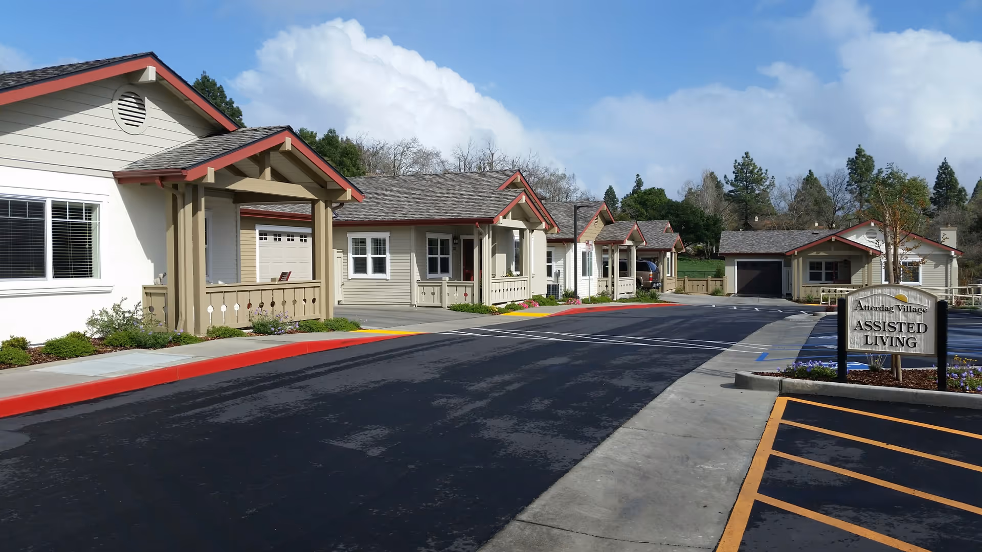 Exterior view of Atterdag Village of Solvang assisted living facility showing a row of single-story residential buildings with beige siding and red trim, a paved driveway, and a sign that reads 'Atterdag Village Assisted Living'.