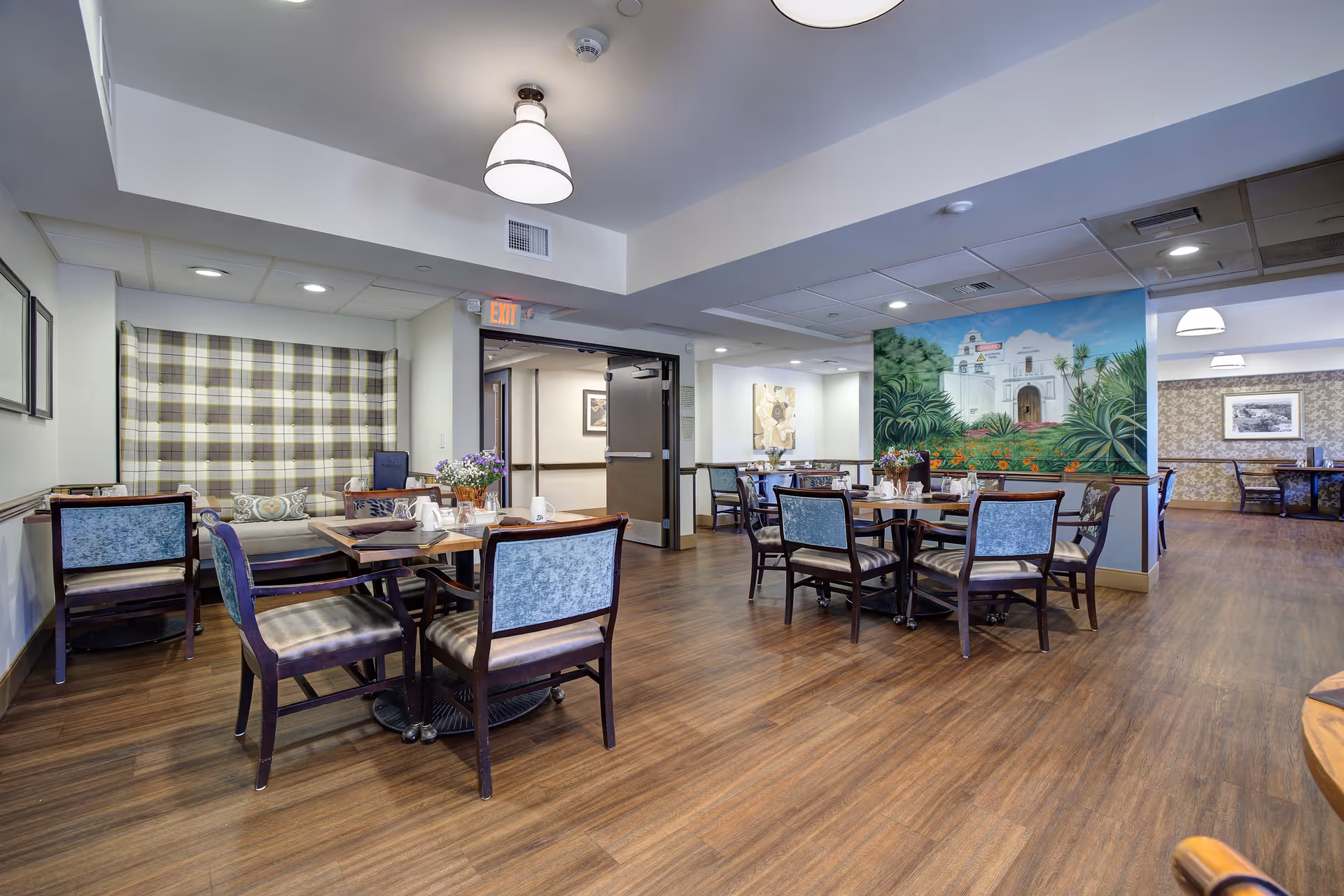 A spacious dining area in a senior living facility with multiple tables and chairs arranged neatly. The room features wood flooring, soft lighting from ceiling fixtures, and a large mural depicting a scenic building with greenery on one wall. There are decorative flowers on the tables and a cozy seating nook with plaid upholstery in the background.