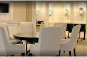 A well-lit interior room with a round table surrounded by six upholstered chairs. In the background, there is a black grand piano and decorative wall art featuring circular designs. The room has a calm and elegant ambiance.