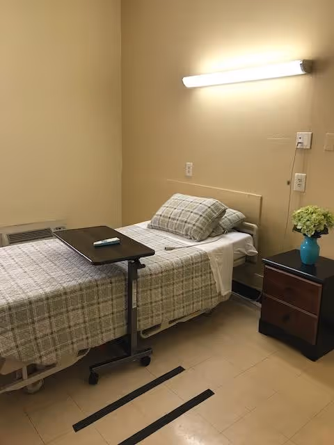 A single hospital-style bed with plaid bedding and a pillow in a small room. There is a rolling overbed table with a remote control on it positioned over the bed. A small dark wooden nightstand with two drawers is next to the bed, holding a blue vase with green flowers. The walls are beige, and a fluorescent light fixture is mounted above the bed. The floor is tiled with beige tiles.