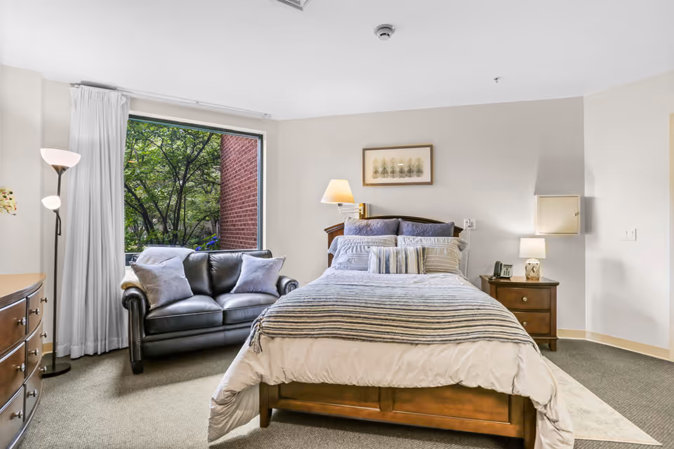 A cozy bedroom with a wooden bed frame, neatly made bed with striped bedding and multiple pillows. To the left of the bed is a black leather loveseat with two gray pillows, positioned in front of a large window showing green trees outside. There is a wooden dresser on the left side of the image and a floor lamp with two lights next to the loveseat. On the right side of the bed is a wooden nightstand with a lamp and a phone. The walls are light-colored and there is a framed artwork above the bed.