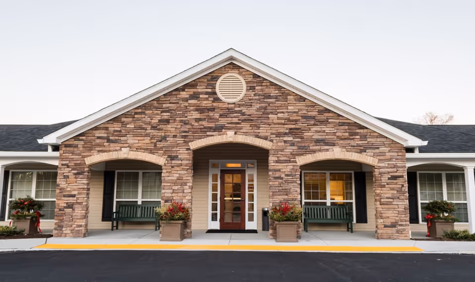 Front exterior view of a senior living facility with a stone facade, a central entrance door with glass panels, two green benches under arched stone coverings, and potted plants with red flowers on either side of the entrance.