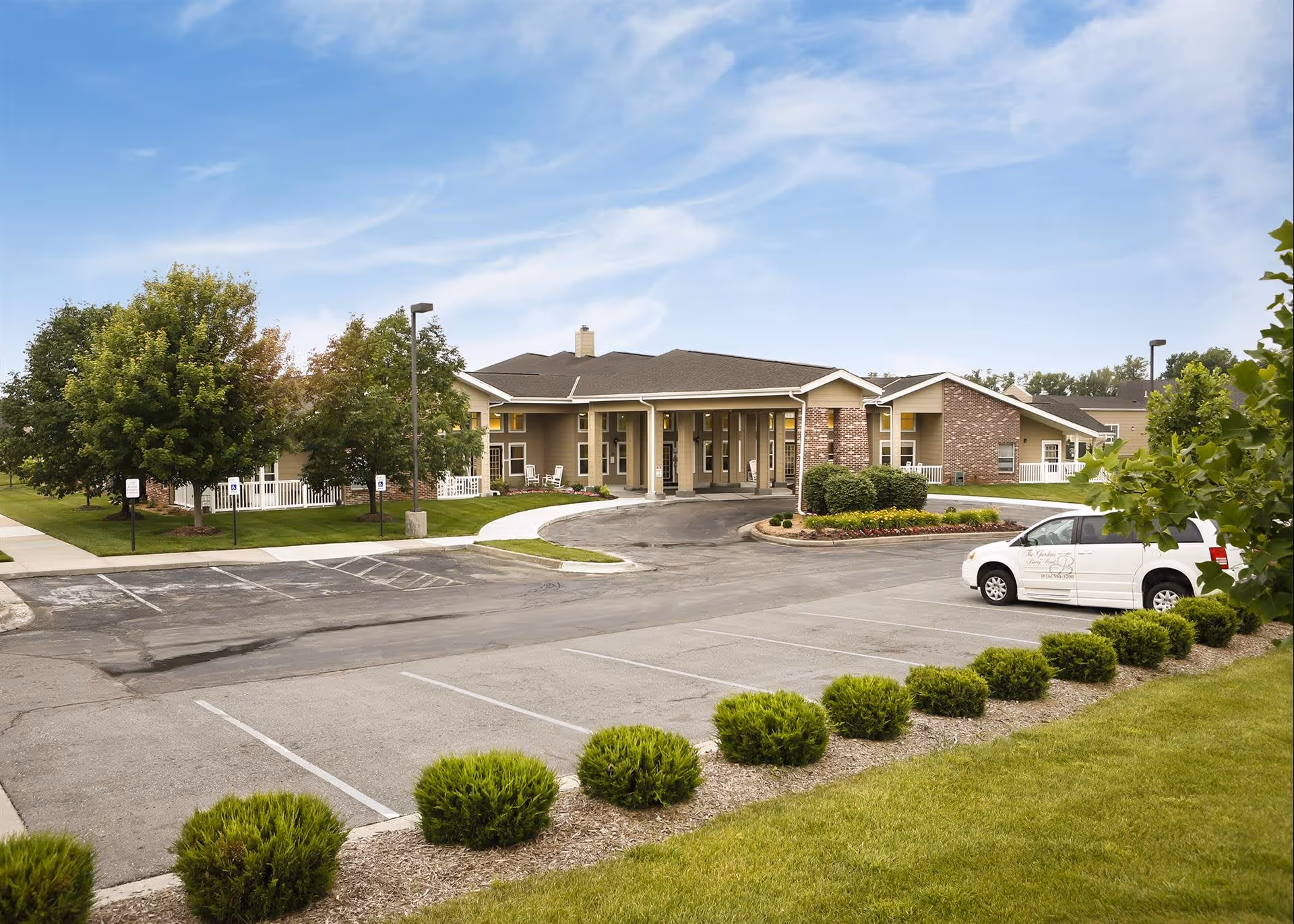 Exterior view of The Gardens at Barry Road Assisted Living and Memory Care building with a parking lot in front, green bushes and trees surrounding the area, and a white vehicle parked on the right side.