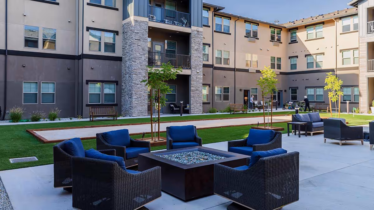 Outdoor courtyard area at Hilltop Springs Senior Living featuring modern wicker chairs with blue cushions arranged around a square fire pit table. The courtyard is surrounded by a multi-story building with balconies and windows. There are small trees planted in the grassy area and benches along the walkway. Several seniors are seen walking and sitting in the background.