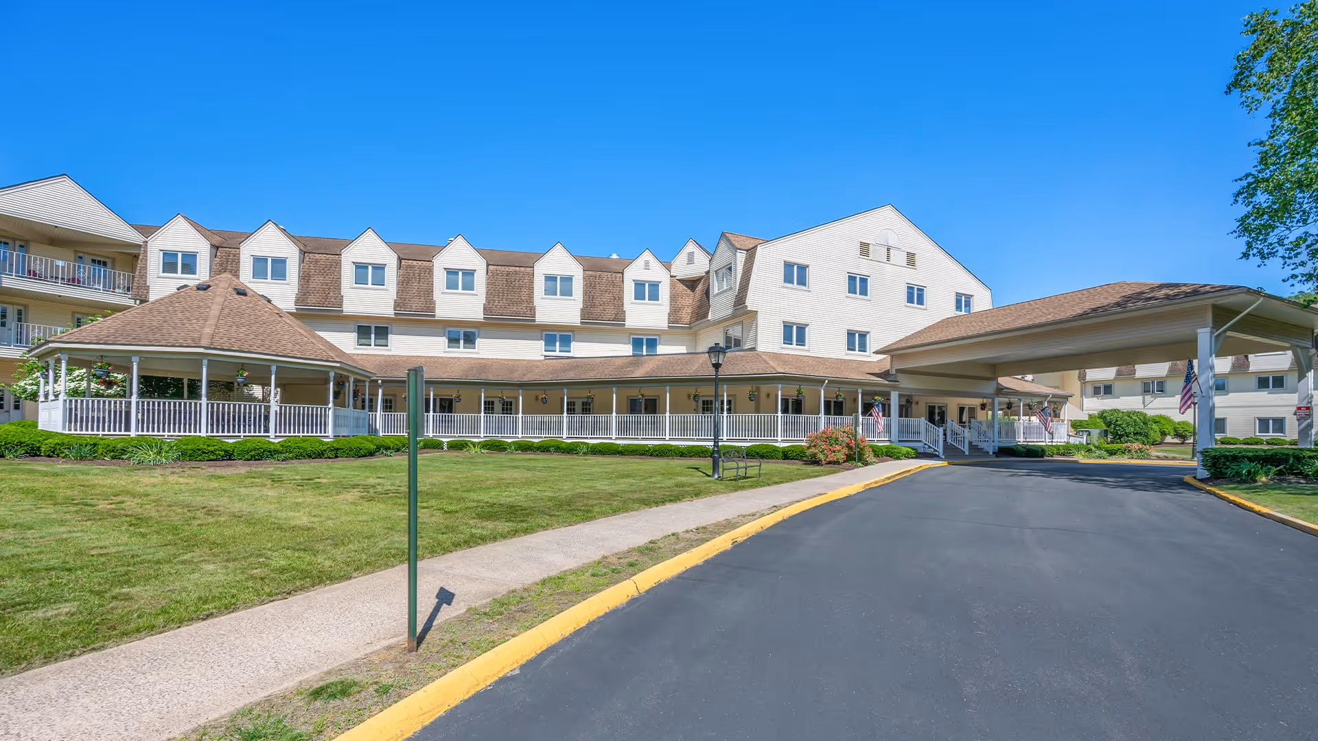 Exterior view of Holiday Farmington senior living facility showing a large multi-story building with beige siding and brown roof shingles. The building features multiple windows and a covered entrance driveway. There is a well-maintained lawn and a paved driveway leading up to the entrance under a clear blue sky.