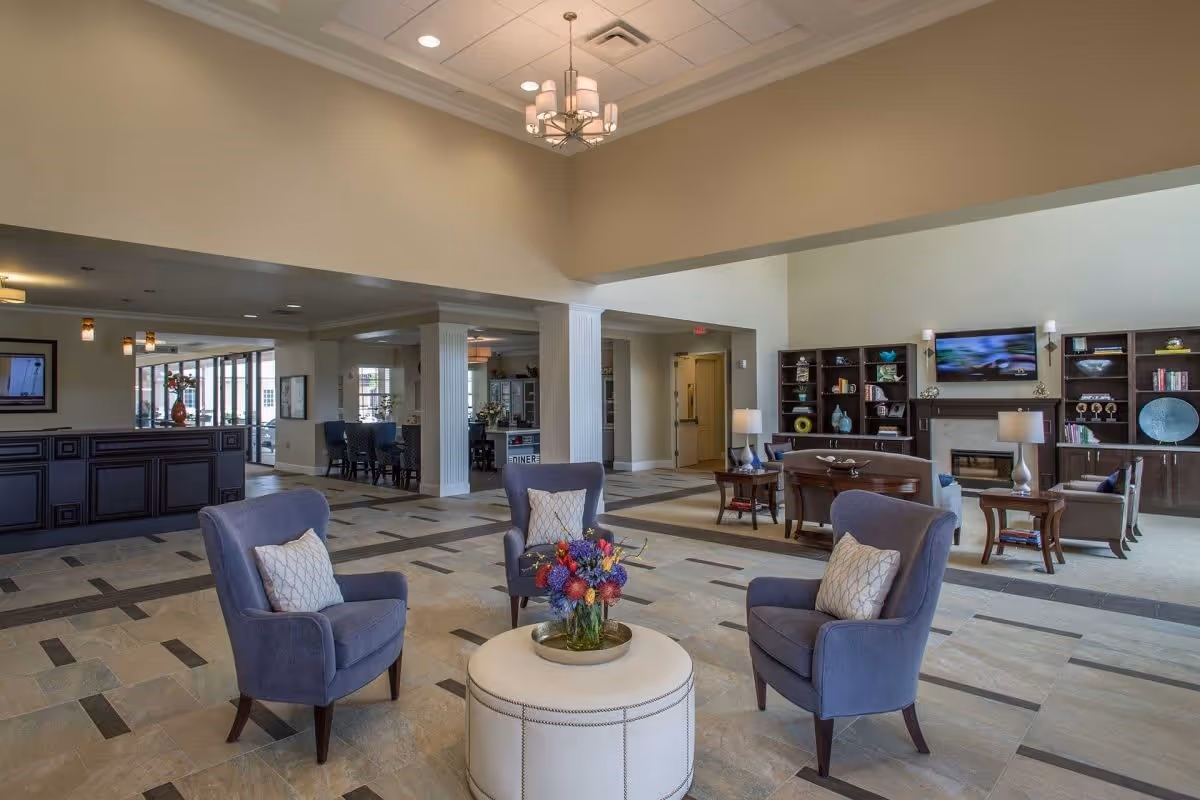 Spacious and well-lit senior living facility common area with four blue armchairs arranged around a round ottoman with a flower arrangement. The room features tiled flooring, a high ceiling with a chandelier, and built-in shelves with decorative items and a mounted TV above a fireplace. In the background, there is a reception desk and a dining area with tables and chairs.