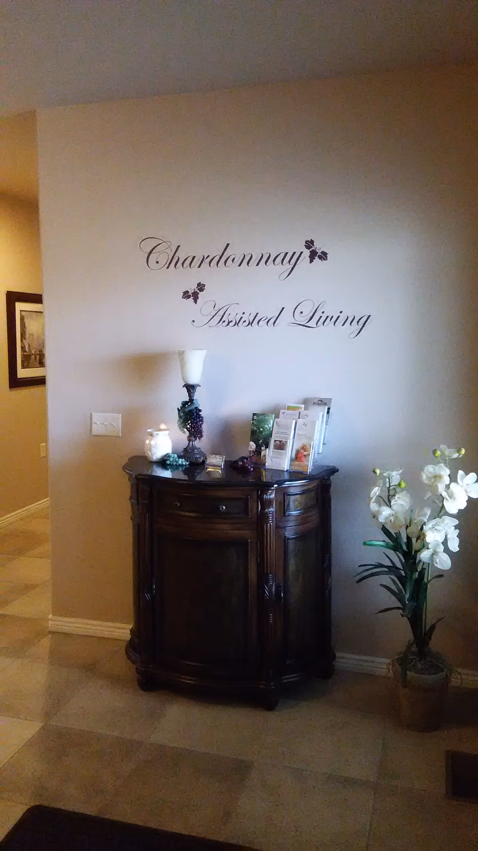 Interior corner of a room with a dark wooden cabinet against a beige wall. Above the cabinet, the words 'Chardonnay Assisted Living' are written in elegant script with small grapevine decorations. On the cabinet are a lamp, a candle, decorative grapes, and several brochures. To the right of the cabinet is a potted plant with white flowers.