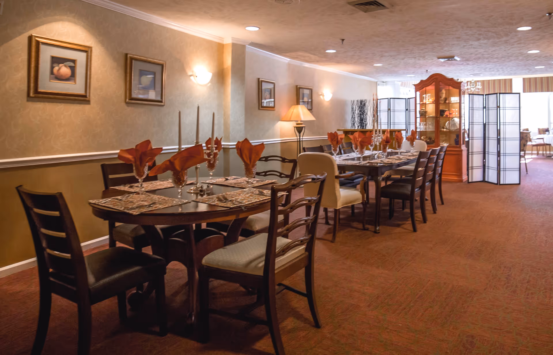 A dining room with two long tables set with placemats, glassware, and folded orange napkins. The room has warm lighting, framed artwork on the walls, a wooden display cabinet, and a folding screen near the windows in the background.