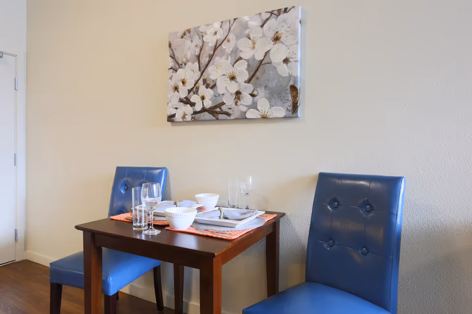 A small dining area with a wooden table set for two, featuring white bowls, plates, glasses, and silverware on orange placemats. Two blue cushioned chairs are positioned on either side of the table. A painting of white flowers on branches hangs on the beige wall above the table.