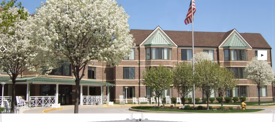 Front exterior of a three-story brick assisted living building with blooming trees, a flagpole, and a covered entrance.