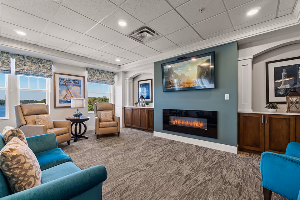 Bright senior living lounge with armchairs and teal sofas, a wall-mounted TV above an electric fireplace, and windows with a water view.