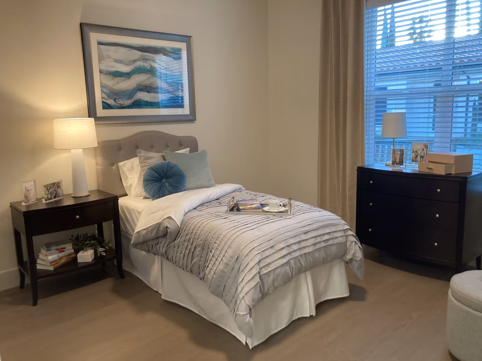 A neatly made single bed with a tufted headboard in a bedroom. The bed has white sheets, a light gray textured comforter, and decorative pillows including a round blue one. A tray with a cup and book is placed on the bed. To the left of the bed is a dark wooden nightstand with a lamp, framed photos, and books on the lower shelf. On the right side of the room is a dark wooden dresser with a lamp, framed photos, and a box on top. A large window with blinds and beige curtains is behind the dresser. A framed abstract artwork hangs on the wall above the bed.