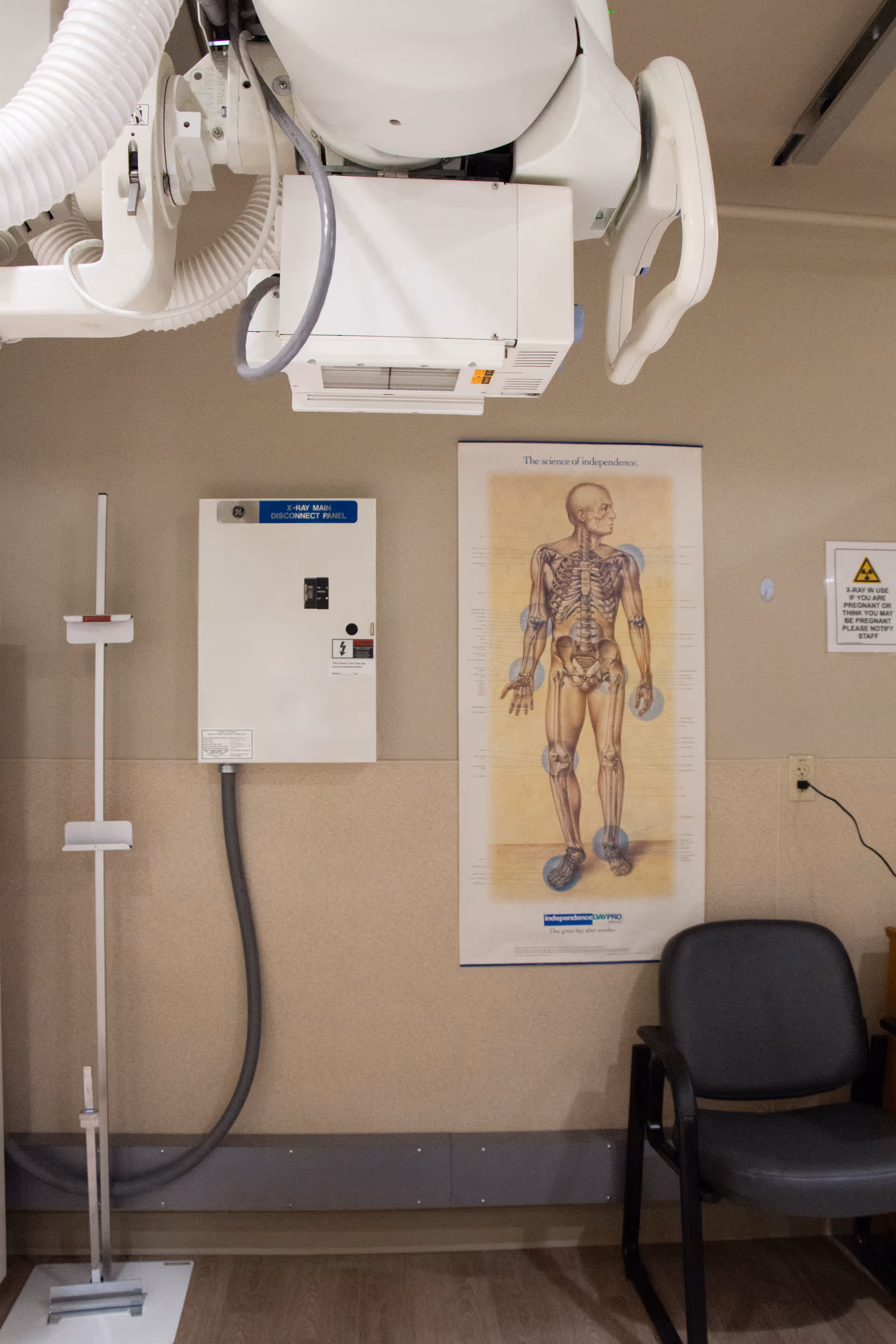 Medical room with an X-ray machine mounted on the ceiling, an X-ray main disconnect panel on the wall, a height measuring device, a chair, and a poster showing a human skeletal system labeled 'The science of independence.'