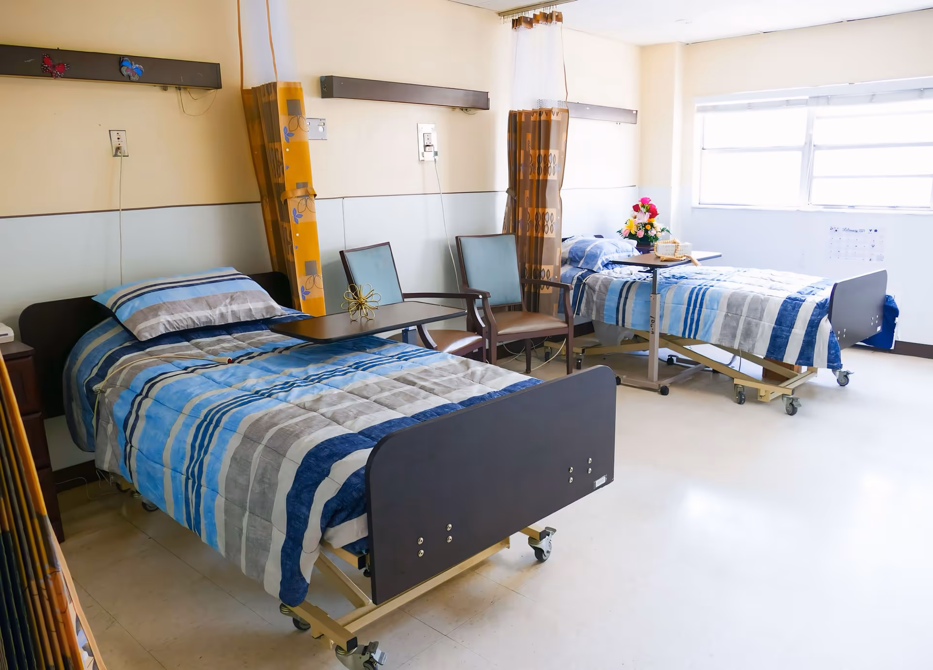 A bright room with two hospital-style beds, each with blue and gray striped bedding. Between the beds are two chairs and a small table with a flower arrangement. The room has large windows allowing natural light to enter, and privacy curtains are partially visible.
