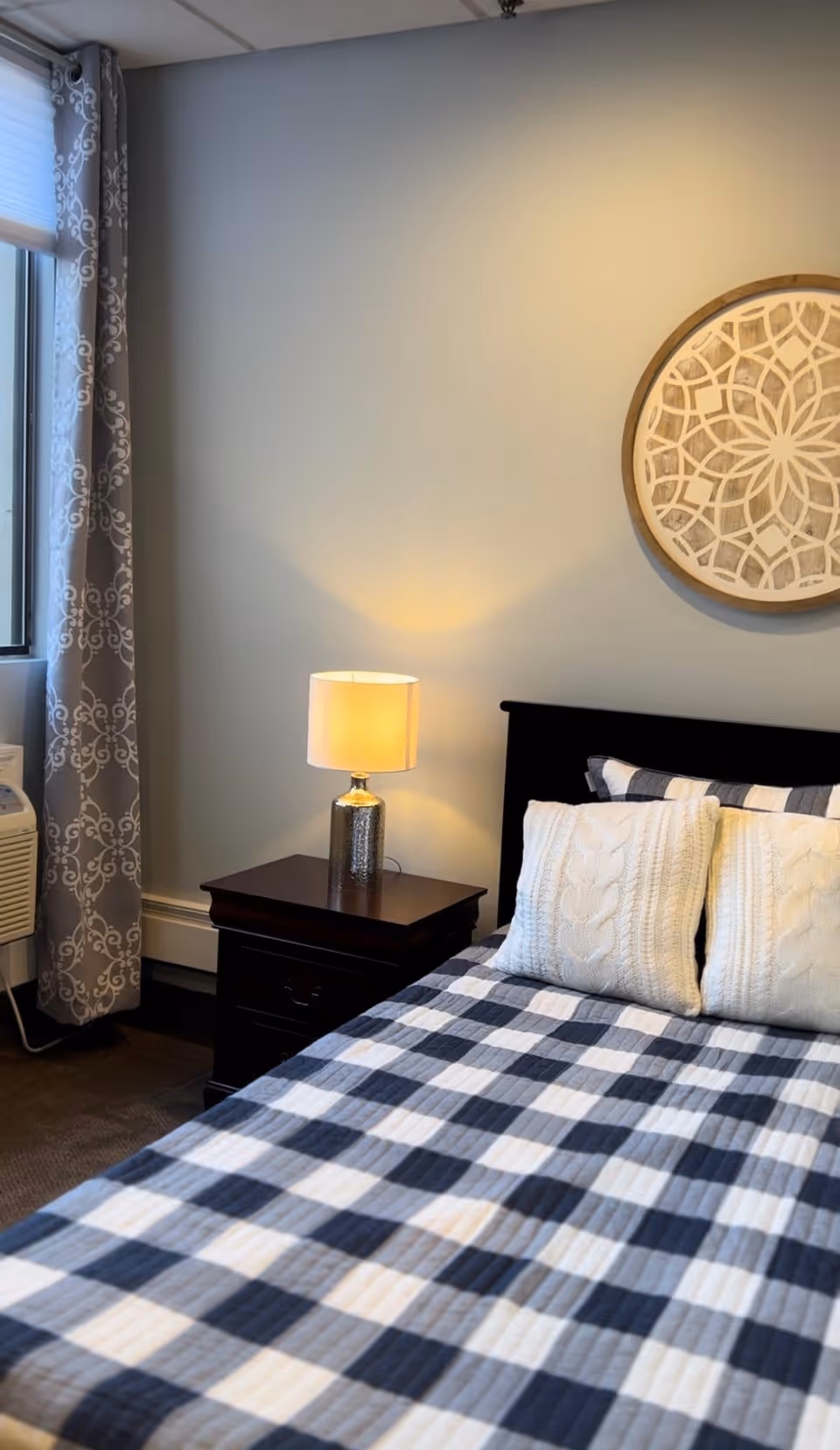 A cozy bedroom featuring a bed with a black and white checkered quilt and two white knitted pillows. Next to the bed is a dark wooden nightstand with a lit table lamp. A decorative round wall art piece hangs above the bed. A window with patterned curtains and an air conditioning unit is visible on the left side.