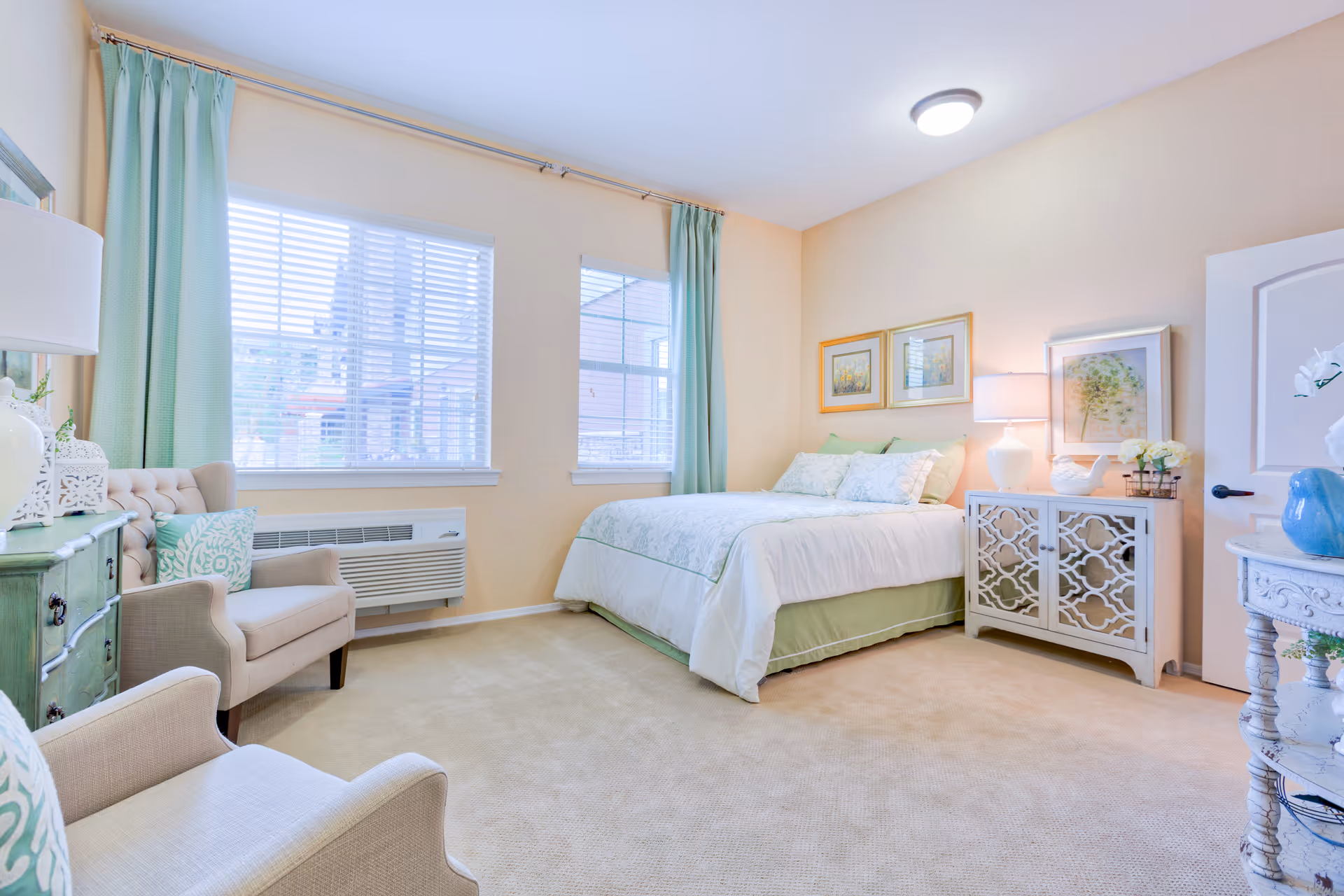 A bright and cozy bedroom in an assisted living facility with a neatly made bed featuring white and green bedding. The room has beige walls and carpet, two windows with light green curtains, two beige armchairs with patterned cushions, a green dresser, and a white nightstand with decorative items and a lamp. There are framed pictures on the wall above the bed and a round white table with a blue vase and flowers near the door.