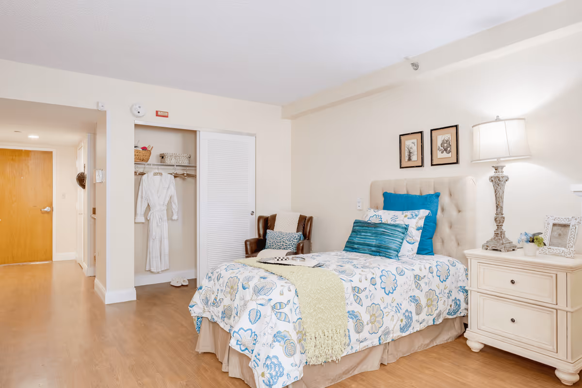 A bright furnished bedroom featuring a floral-patterned single bed with pillows and a throw, a nightstand with a lamp, an armchair, and an open closet with a robe.