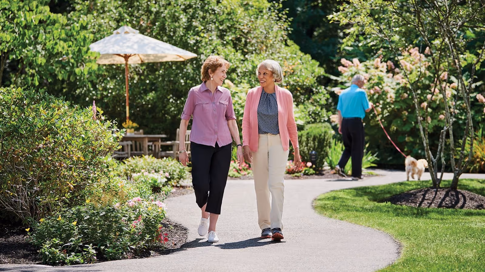 Two elderly women walking and talking on a paved garden path surrounded by lush greenery and flowers. In the background, an elderly man is walking a small dog. There is a patio table with an umbrella visible among the plants.