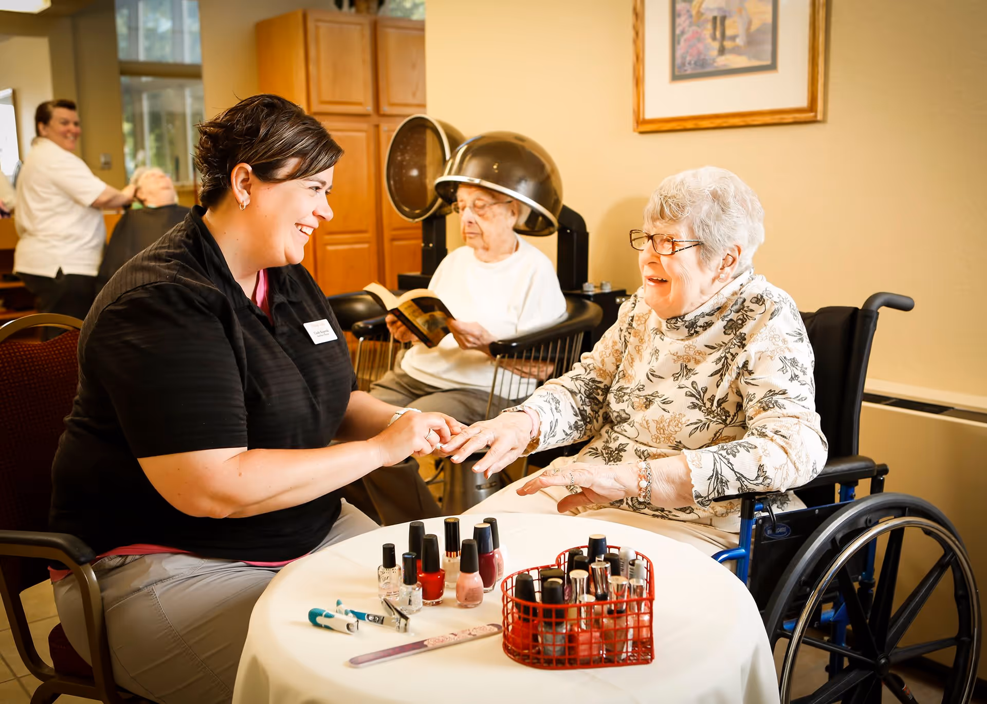 A caregiver is painting the nails of an elderly woman in a wheelchair at a table with various nail polish bottles and manicure tools. In the background, another elderly woman is sitting under a hair dryer reading a book, while a staff member attends to another person.
