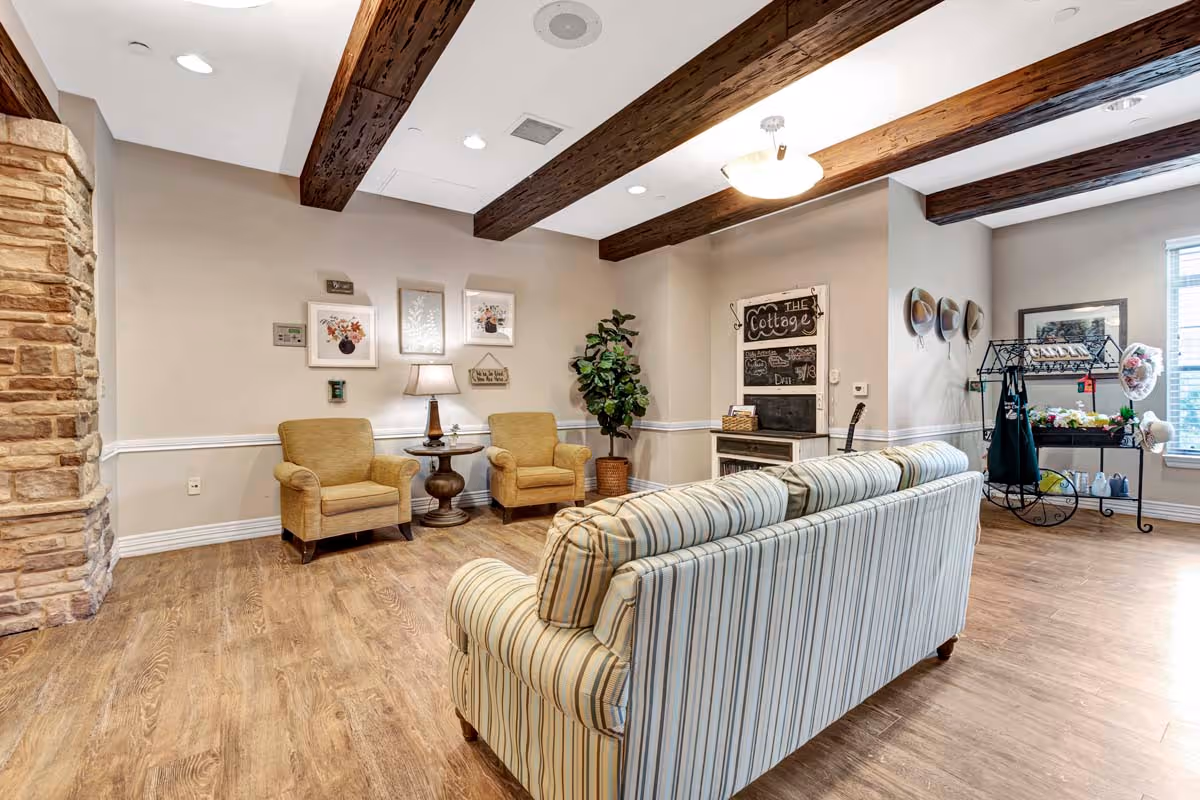 A cozy living room area with wooden ceiling beams, a striped sofa facing away, two beige armchairs with a small round table and lamp between them, framed floral artwork on the wall, a potted plant, and a decorative chalkboard labeled 'The Cottage'. There are hats hanging on the wall and a small cart with various items near a window letting in natural light.