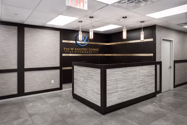 Reception area of The W Assisted Living at Riverdale featuring a modern front desk with gray and dark wood paneling, three pendant lights hanging from the ceiling, and a sign on the wall displaying the facility's name and logo.