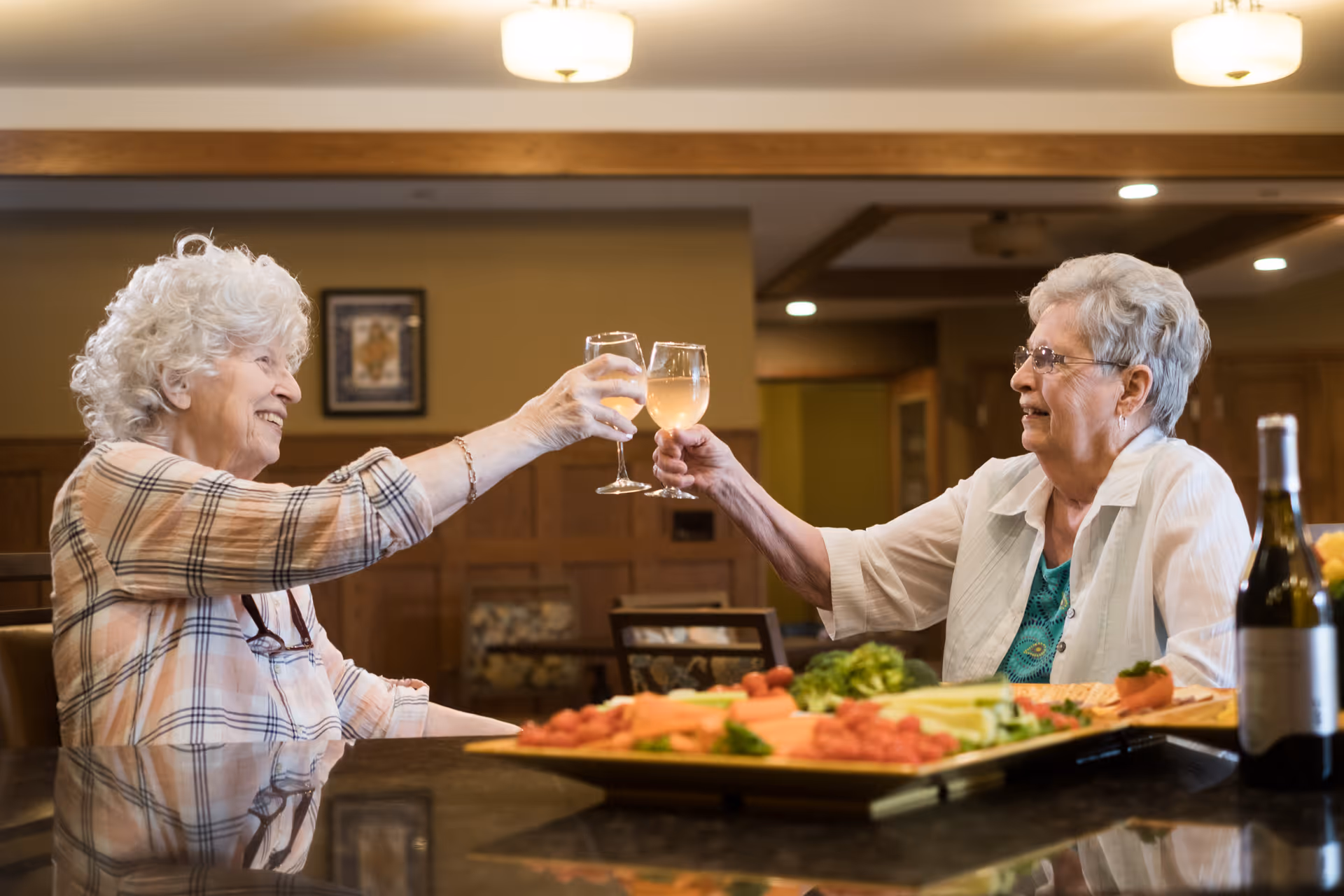 Two elderly women sitting at a table in a dining area, smiling and clinking glasses of white wine. The table has a platter of assorted vegetables and meats, and a bottle of wine is visible. The background shows warm lighting and wooden paneling.