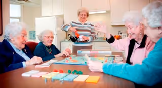 A group of elderly women sitting around a table playing a board game together in a bright room with a kitchen area in the background. One woman is standing and pouring a drink while the others are seated and engaged in the game, smiling and interacting.