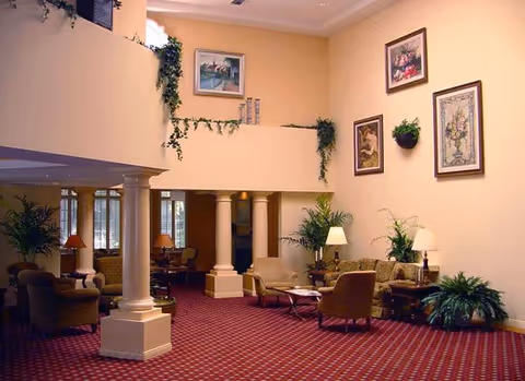 Spacious living room area with high ceilings, beige walls adorned with framed artwork and hanging plants, red patterned carpet, multiple seating arrangements including armchairs and sofas, side tables with lamps, and large white columns separating the space.