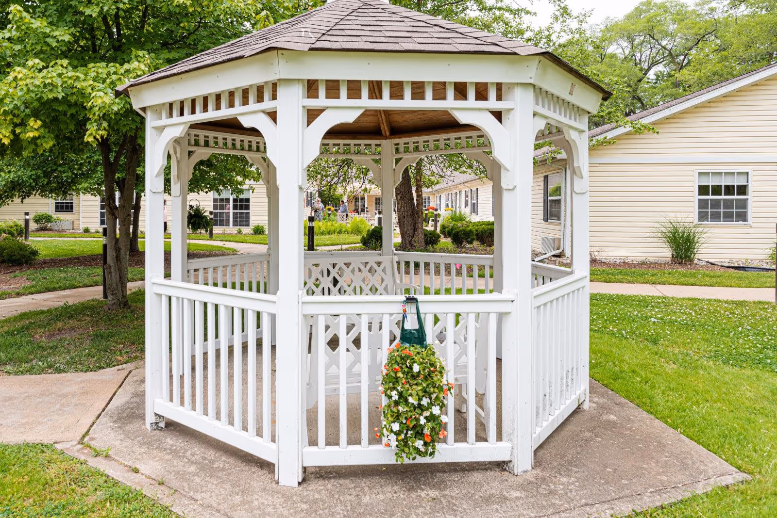 A white wooden gazebo with a shingled roof situated on a concrete pad in a grassy outdoor area. There is a hanging flower basket with green leaves and white and orange flowers attached to the gazebo railing. Surrounding the gazebo are trees, shrubs, and beige buildings with windows, indicating a landscaped community setting.