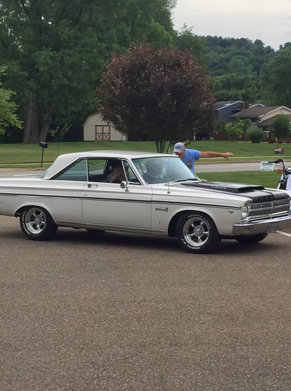 A white vintage two-door car parked on pavement with a person leaning into the driver's window and trees and houses behind.