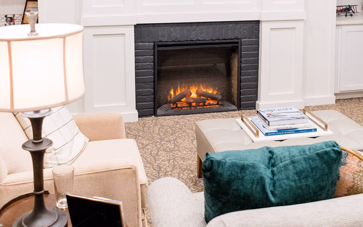 Cozy living room with a lit fireplace, upholstered chairs, a tufted ottoman with books, and a floor lamp.