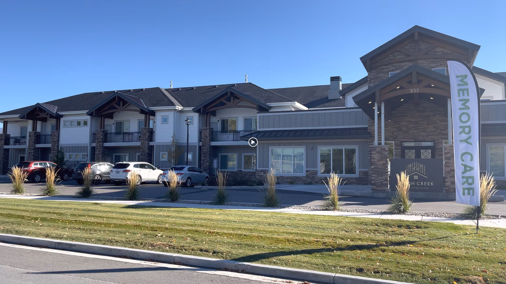 Exterior view of Birch Creek Assisted Living of Smithfield, a two-story building with stone and wood accents. Several cars are parked in front, and a tall flag with the words MEMORY CARE is displayed near the entrance under a covered porch.