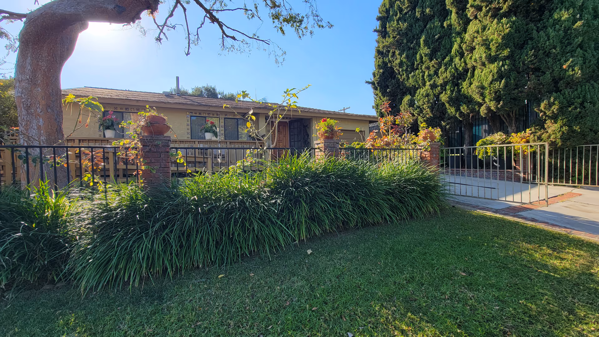 Exterior view of a single-story building with a front yard featuring green grass, bushes, and a large tree. The building has a porch with potted plants and a railing, and there is a paved walkway leading to the entrance. Tall trees are visible on the right side under a clear blue sky.