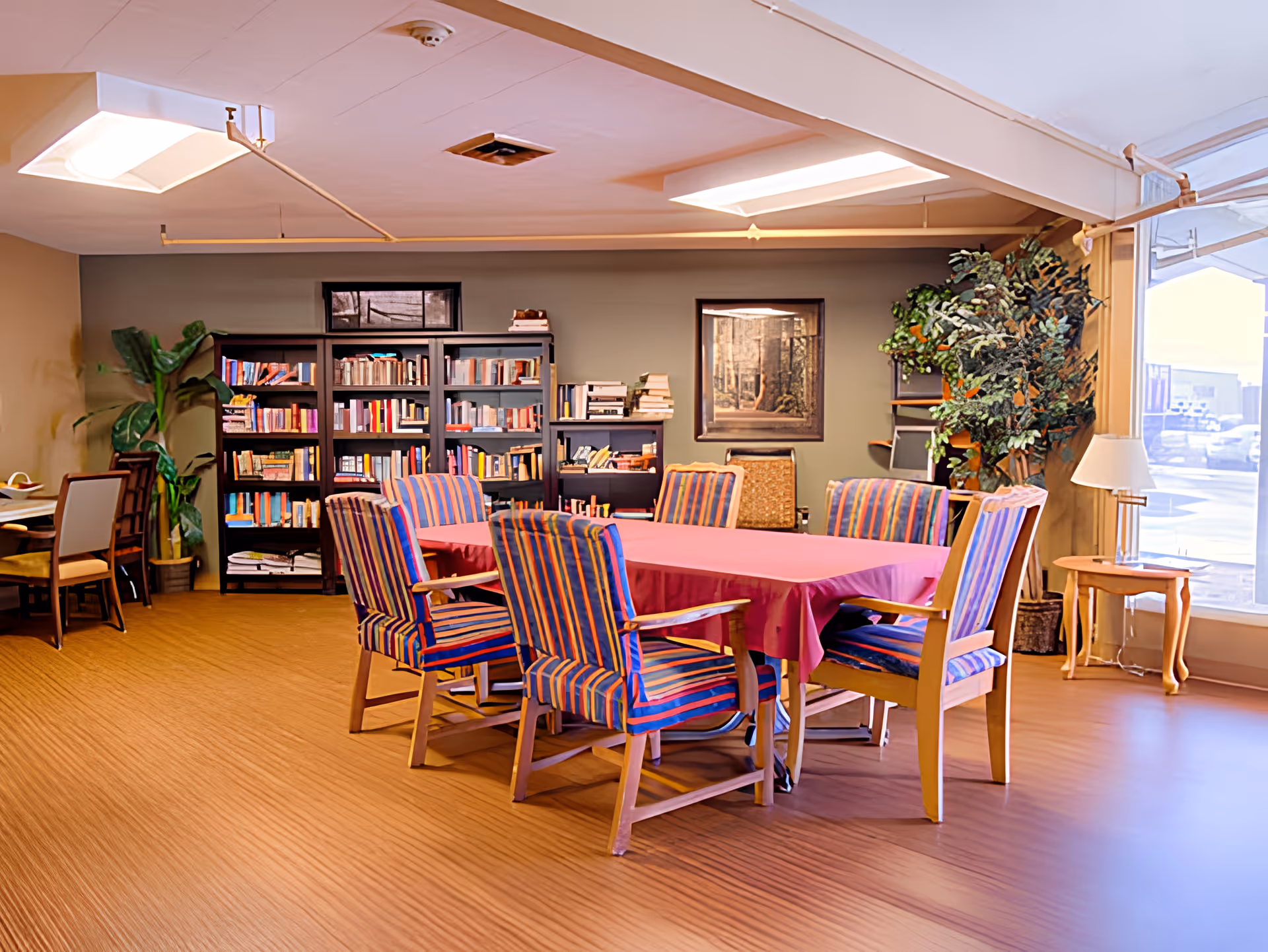 A well-lit room with a rectangular table covered with a pink tablecloth surrounded by six wooden chairs with striped upholstery. Behind the table, there are bookshelves filled with books and a framed picture on the wall. A large potted plant is in the corner near a window with a small wooden side table and a lamp beside it. The floor has a wood-like texture and the ceiling has recessed lighting.