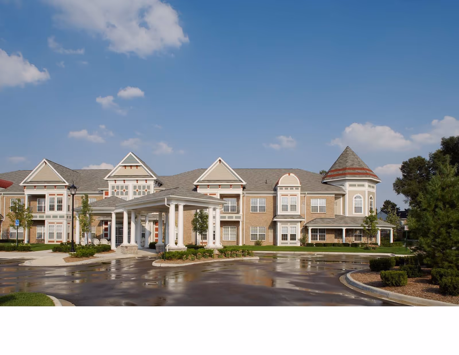 Front exterior of a two-story senior living building with a covered porte-cochère, turret, and wet circular driveway under a blue sky.