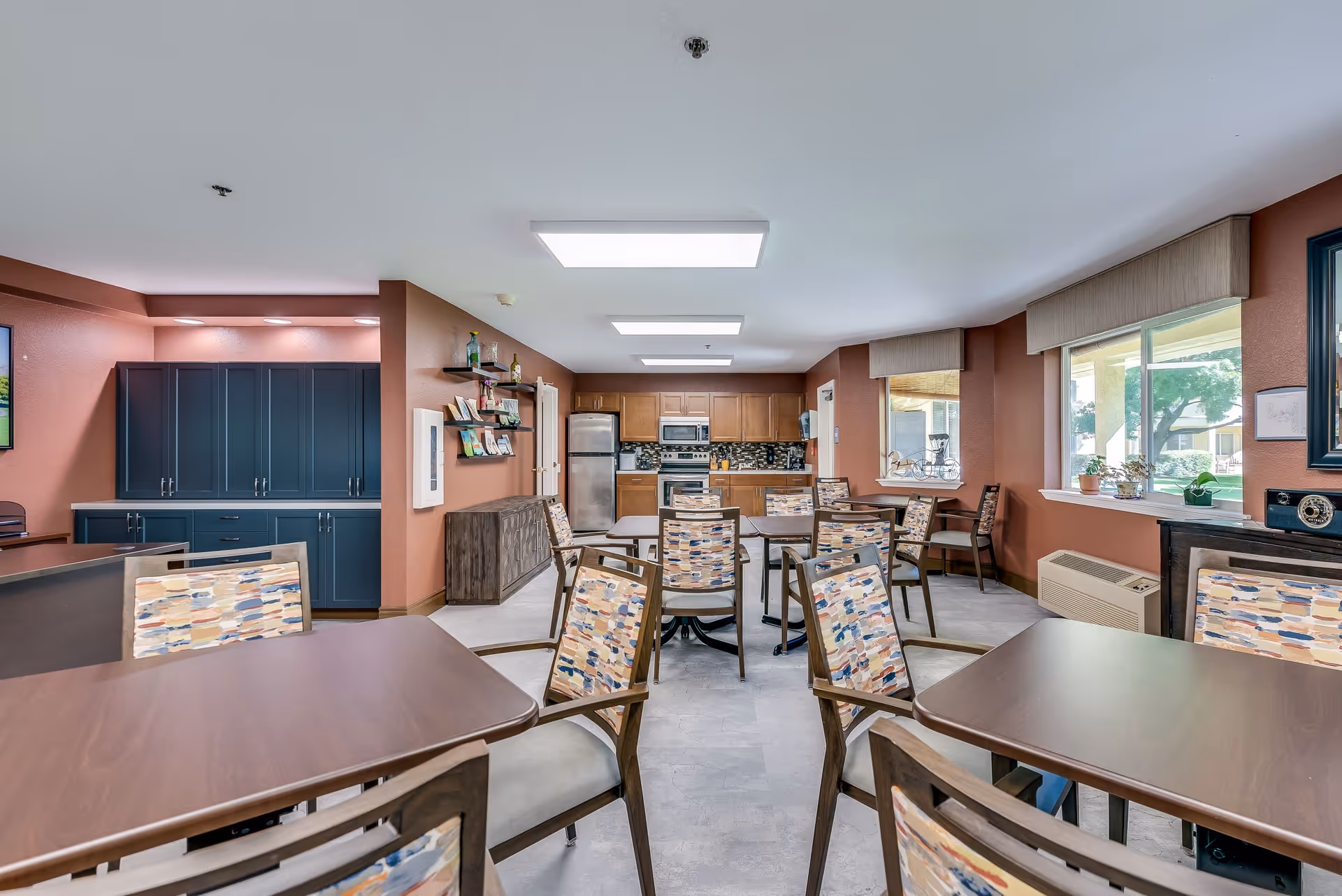 A bright and spacious dining area in a senior living facility featuring multiple wooden tables and chairs with patterned cushions. The room has large windows with valances, allowing natural light to fill the space. In the background, there is a kitchen area with stainless steel appliances, wooden cabinets, and a tiled backsplash. The walls are painted in warm tones, and there are shelves with decorative items and plants on the windowsills.