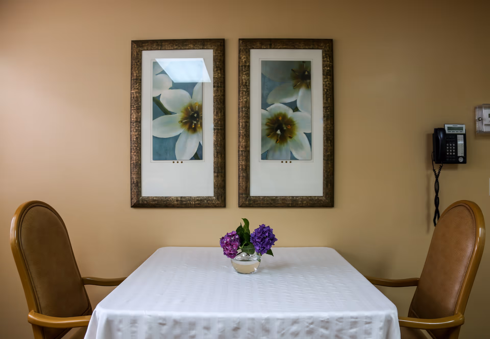 A small table covered with a white tablecloth with a small glass vase holding purple flowers in the center. Two brown cushioned chairs with wooden armrests are positioned on either side of the table. On the beige wall behind the table, there are two framed pictures of white flowers with green backgrounds. A black wall-mounted telephone is visible on the right side of the wall.