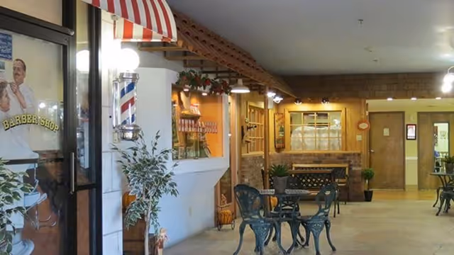 Indoor assisted-living common area featuring small metal cafe tables and chairs, a barber shop storefront with a striped awning, and wood-paneled decorative walls.