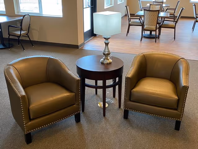 Two brown leather armchairs with nailhead trim are positioned on either side of a round wooden side table with a silver lamp on top. In the background, there are additional chairs and tables near large windows letting in natural light.