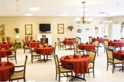 A spacious dining room with multiple round tables covered with red tablecloths, each surrounded by wooden chairs. The room has light-colored walls decorated with framed artwork and plants. A flat-screen TV is mounted above a white fireplace, and a chandelier hangs from the ceiling. Large windows and glass doors allow natural light to enter the room.