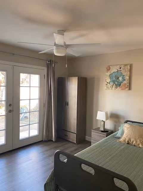 A bedroom with a single bed covered in a green quilt and a beige pillow. Next to the bed is a small nightstand with a lamp. A wooden wardrobe stands against the wall, and above it hangs a floral painting with a large blue flower. There are double glass doors with beige curtains leading outside, and a white ceiling fan is mounted on the ceiling.