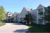 Three-story light-blue senior living building with balconies, trees, and a curved driveway.