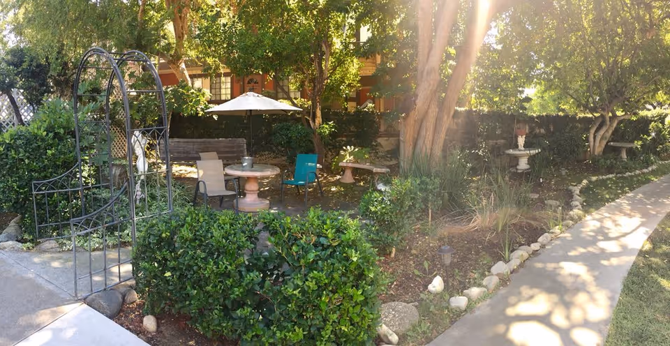 Shaded courtyard with a patio table, chairs and umbrella, an arched trellis, benches, trees and a curved walkway.
