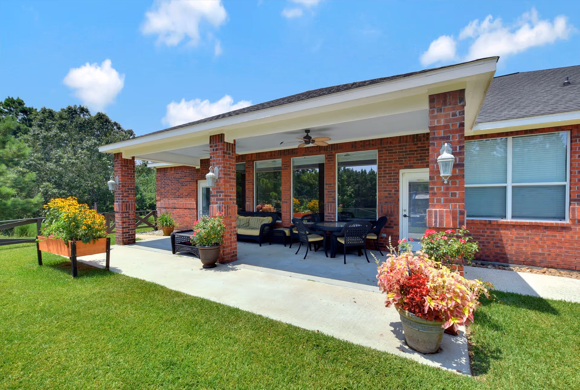 Outdoor covered patio area attached to a red brick building with a ceiling fan, outdoor seating including a sofa and chairs around a table, potted plants, and a well-maintained grassy lawn with flower beds under a bright blue sky with some clouds.